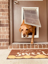 A golden labrador dog is halfway through a dog door on a brown door with a brick exterior, above a mat that says