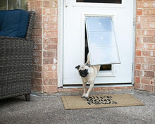 A pug walks through a pet door with a doormat that reads