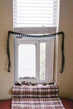 Window with closed blinds and a pom-pom garland, decorated with a patterned throw.