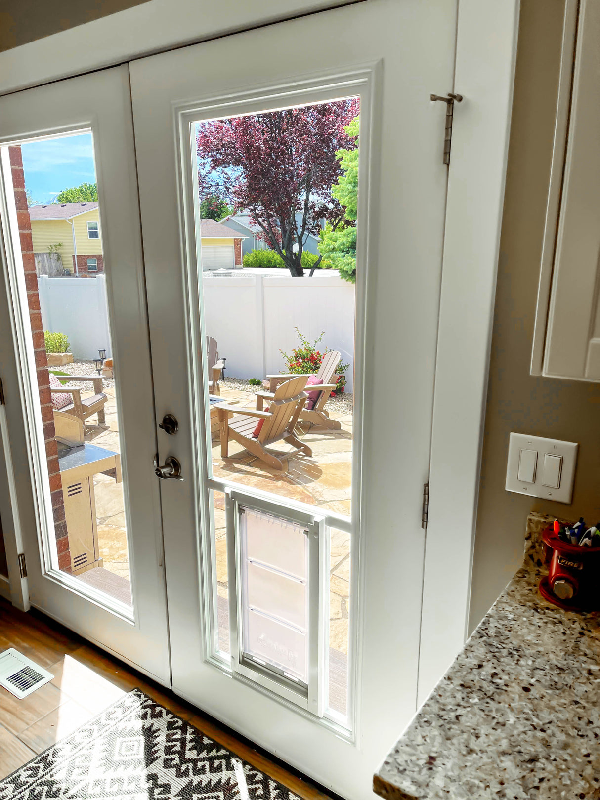 Interior view of white patio doors with a dog door insert, leading to a backyard patio with Adirondack chairs.