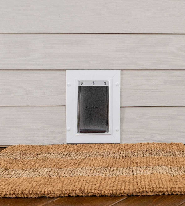 A pet door is installed in light siding above a natural fiber mat.