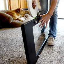 A person applies double-sided tape to a black-framed glass panel, with a dog resting on a nearby couch.