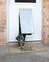 A small, gray dog exits a white door through a pet door onto a "Wipe Your Paws" doormat.
