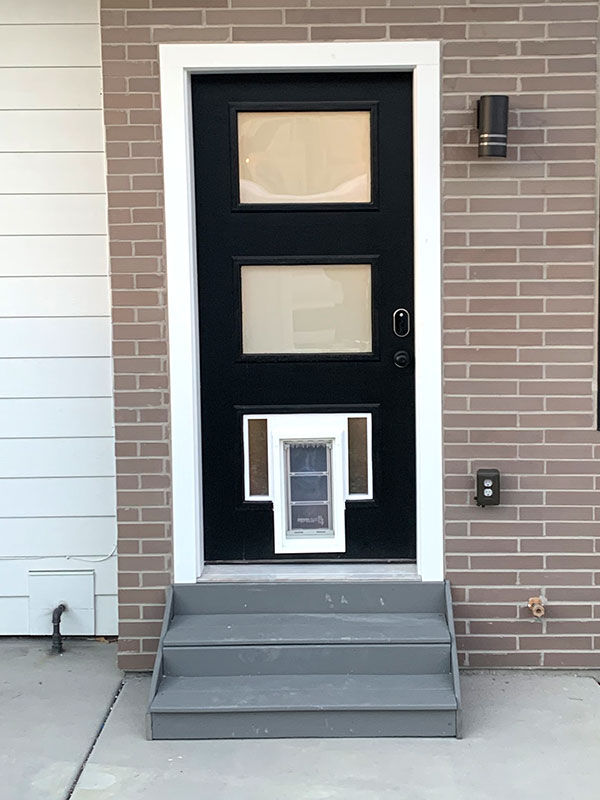 A black door with two windows and a dog door sits atop gray steps in front of a brick and white-sided house.
