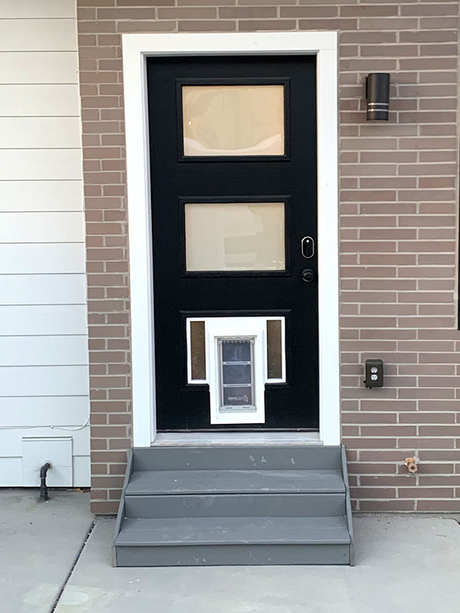 A black door with two windows and a dog door sits atop gray steps in front of a brick and white-sided house.