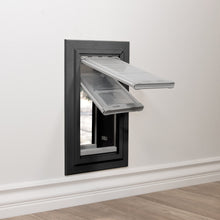 A pet door with clear flaps is installed in a white wall.