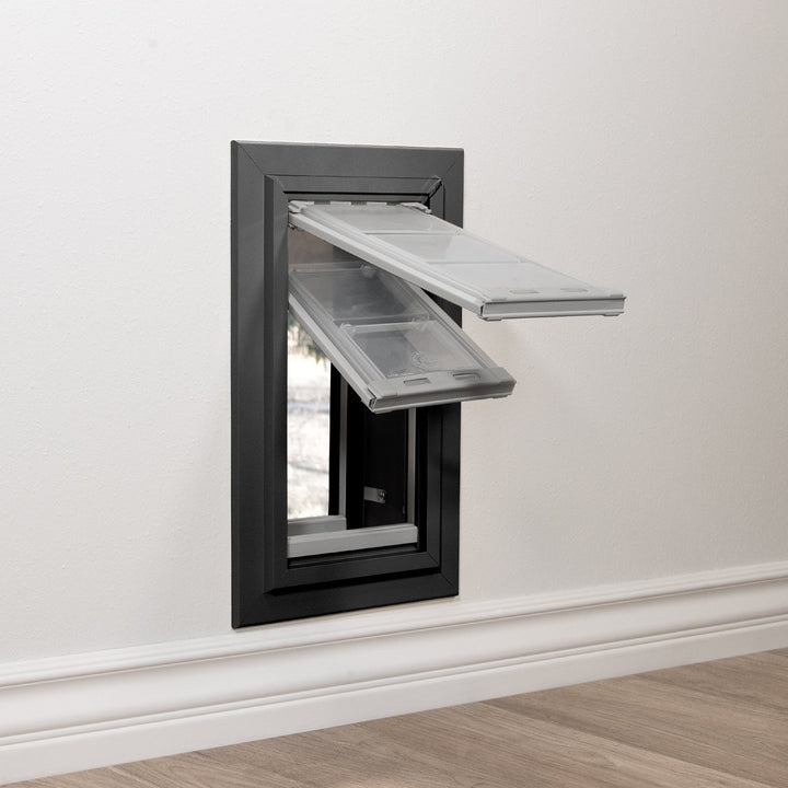 A pet door with clear flaps is installed in a white wall.