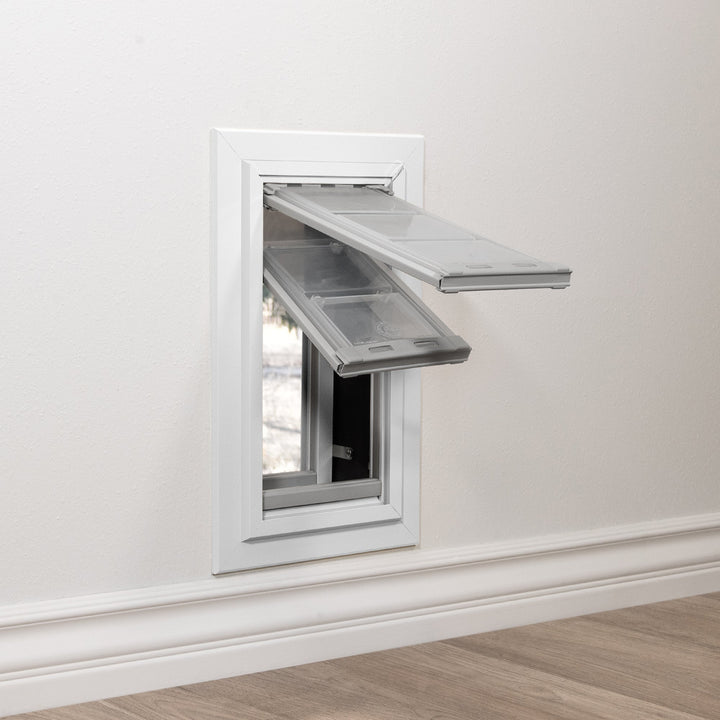 A pet door with two open flaps is installed in a white wall above a baseboard.
