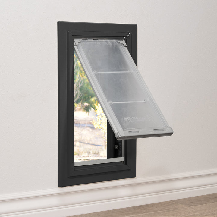 A black-framed pet door is installed in a wall, revealing an outdoor view through its partly-open, flexible flap.
