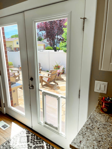 Interior view of white patio doors with a dog door insert, leading to a backyard patio with Adirondack chairs.