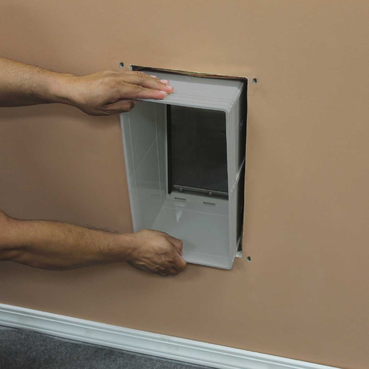 Hands installing a rectangular white plastic pet door into a wall.