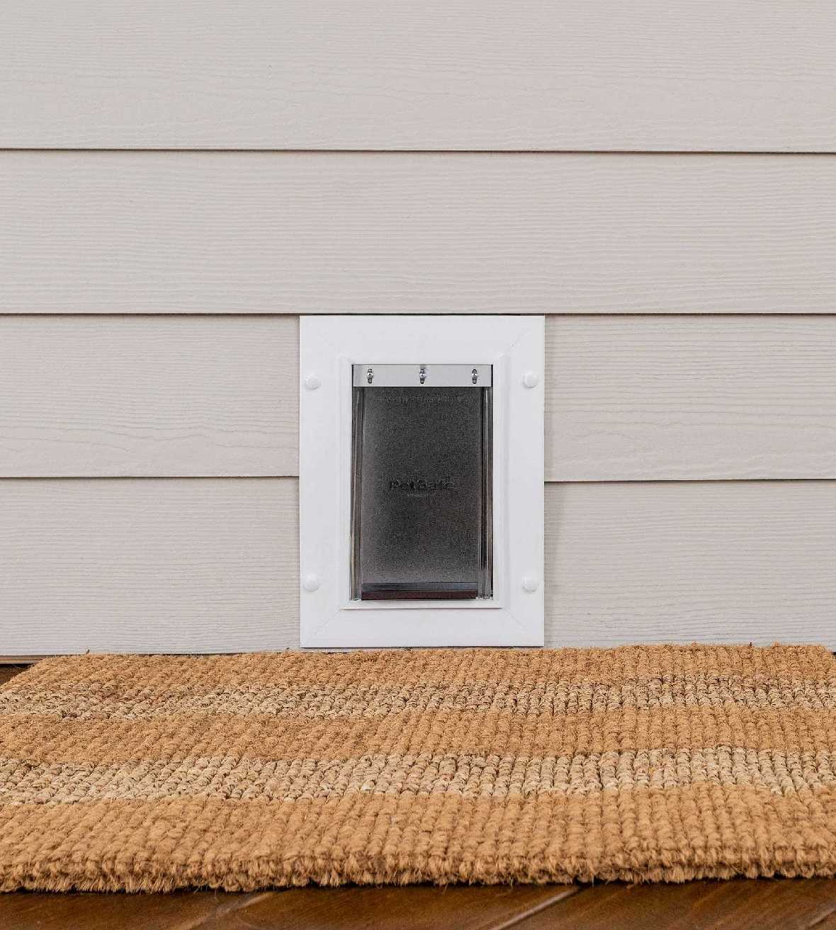 A pet door is installed in light siding above a natural fiber mat.