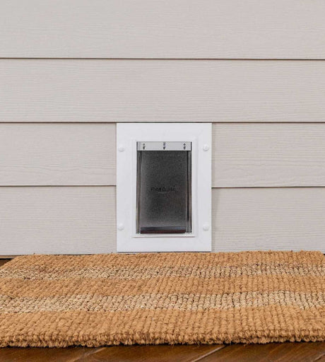 A pet door is installed in light siding above a natural fiber mat.